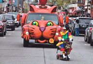 A van resembling an orange cat follows a clown driving a mobility scooter